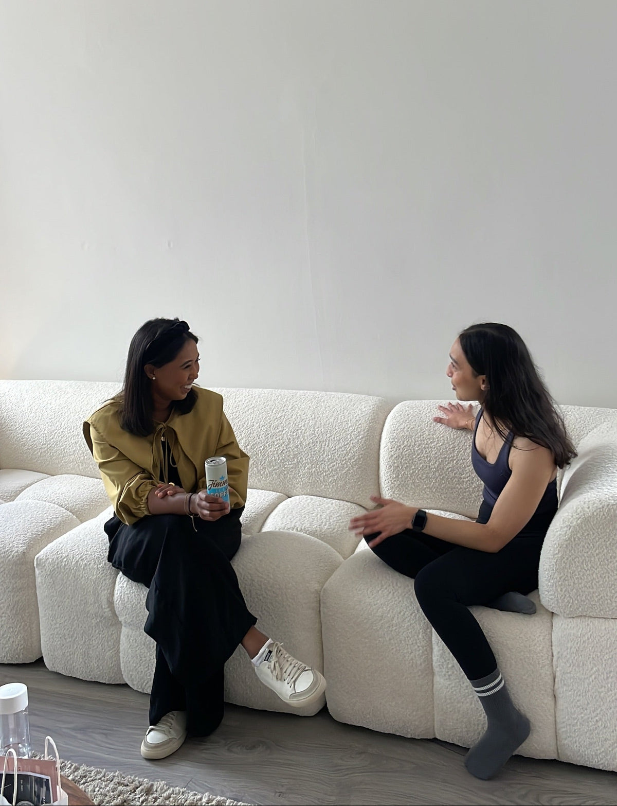 Two women laughing and chatting, sitting on a white couch in a minimalistic room.